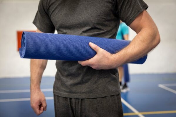 A man in grey prison sweatshirt and joggers holding a purple yoga mat