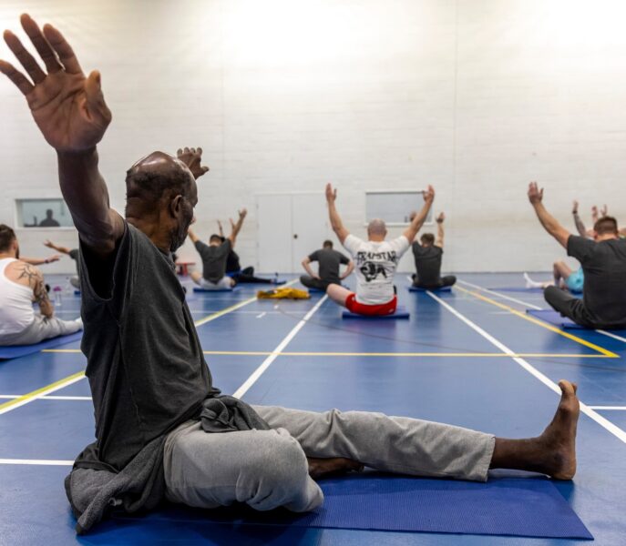 Man sitting on yoga mat in prison gym, arms outstretched, looking away at others taking part in yoga class