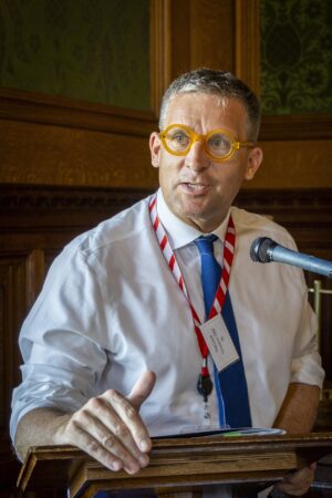 Lord James Timpson in shirt and tie wearing bright spectacles at a lectern in the River Room, House of Lords